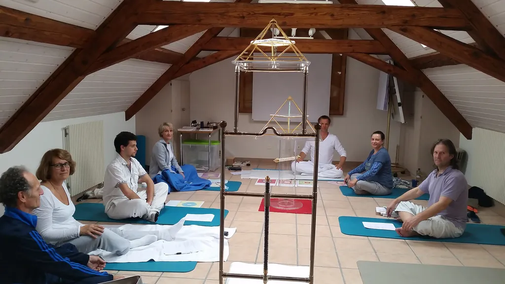 Gruppe von sieben Personen auf Yogamatten in einem Dachgeschossraum mit sichtbaren Holzbalken; im Vordergrund eine Horus Energiepyramide auf einem Metallständer, an der Decke eine weitere goldene Pyramidenstruktur.