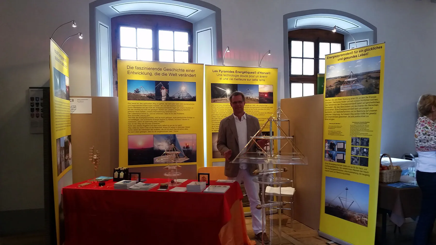 Mats Björkman steht hinter einem rot gedeckten Ausstellungstisch mit Horus-Energiepyramiden-Modellen aus Acrylglas; im Hintergrund gelbe Rollup-Banner mit deutschen und französischen Texten über die Pyramiden, in einem Saal mit gewölbten Fenstern.