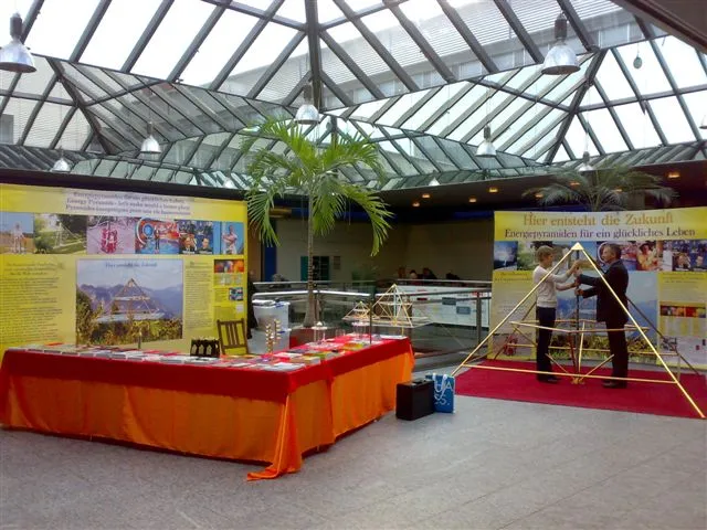 Ausstellungsstand mit orangerot gedecktem Tisch, Horus-Energiepyramiden-Modellen und Infomaterialien; im Hintergrund bauen zwei Personen eine grosse Pyramidenkonstruktion aus Metallstäben auf, flankiert von gelben Bannern mit dem Text Hier entsteht die Zukunft – Energiepyramiden für ein glückliches Leben