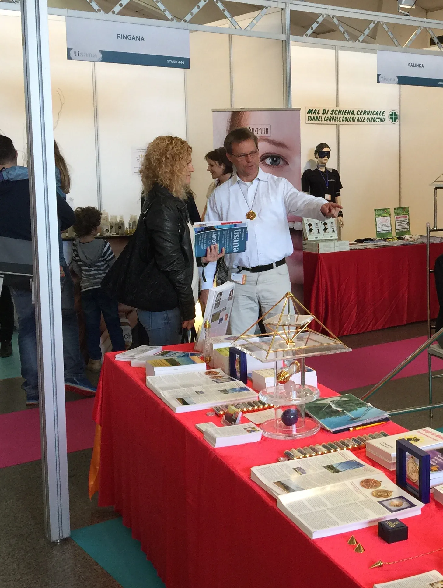 Mats Björkman im Gespräch mit einer Besucherin an einem Messestand mit einer goldenen Horus Energiepyramide, Büchern und Informationsmaterialien auf einem rot bedeckten Tisch, Messehallenumgebung.