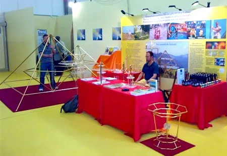 Messestand mit goldenen Horus-Energiepyramiden auf rot gedeckten Tischen sowie einer großen Pyramiden-Gestell-Skulptur im Vordergrund; eine Person steht am Stand in einer Messehalle.