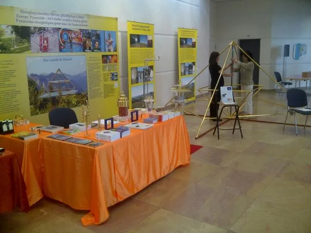 Messestand mit orangem Tischtuch, Büchern und Ausstellungspostern; im Hintergrund wird ein grosses goldenes Pyramidengestell aufgebaut.