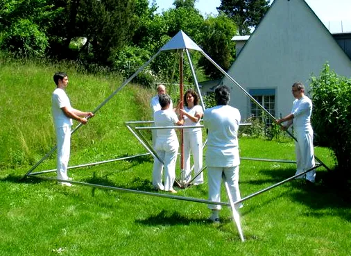 Gruppe von Personen in weißer Kleidung steht auf einer Wiese um ein aufgebautes Metallgestell einer Horus Energiepyramide, im Hintergrund ein Gebäude und Bäume.