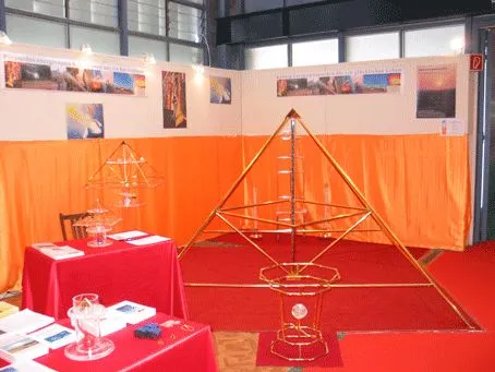 Messestand mit einer großen goldenen Horus-Energiepyramide vor orangem Vorhang, rot gedeckten Tischen mit Materialien und Infopostern.