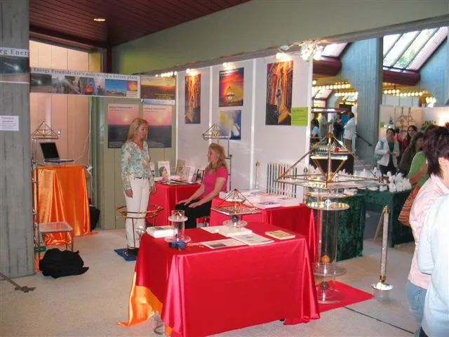 Messestand mit zwei Frauen, rot und orange gedeckten Tischen, goldenen Energiepyramiden und Kristallsäulen; Besucher und Infoposter im Hintergrund.