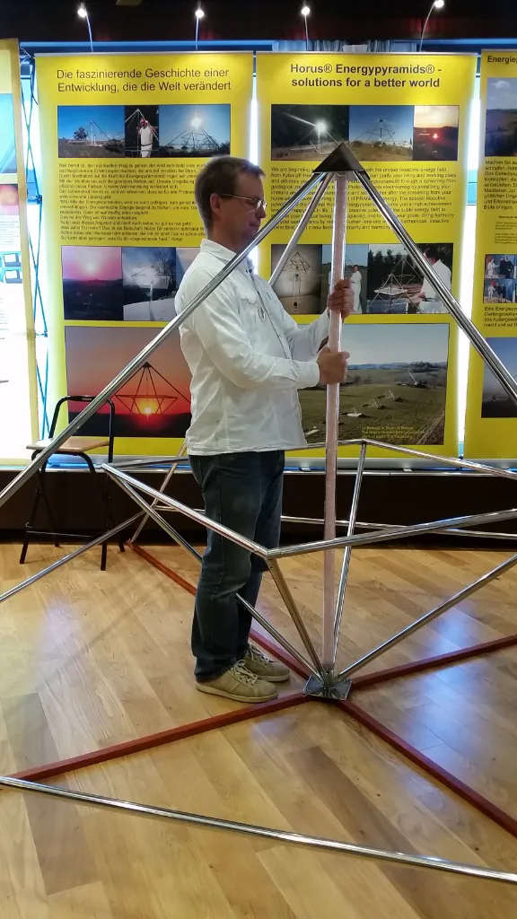 Mats Björkman hält die zentrale Achse eines grossen Horus Energiepyramiden-Modells aus Metallstäben vor gelben Messebanners mit dem Schriftzug "Horus Energypyramids – solutions for a better world".