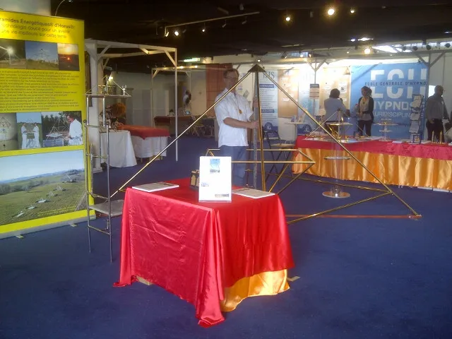 Messestand mit einer großen Horus Energiepyramide aus Metallstäben und rot-goldenem Tisch im Ausstellungsraum; links ein gelbes Roll-up-Banner mit Fotos der Pyramiden, im Hintergrund weitere Messestände.