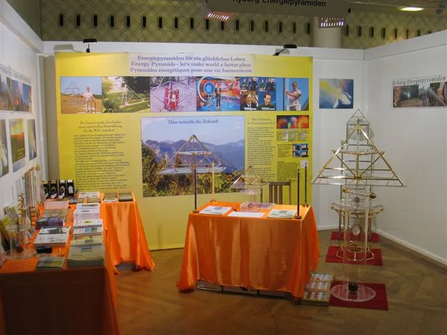 Messestand mit orange gedeckten Tischen, einer großen goldenen Horus-Energiepyramide und gelben Infowänden zum Thema Energiepyramiden.