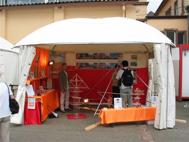 Messestand unter weissem Zeltdach mit orangefarbenen Tischdecken, auf denen mehrere Horus Energiepyramiden-Modelle ausgestellt sind, und zwei Personen im Stand.