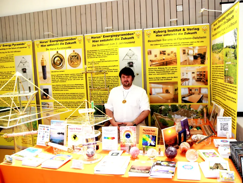 Tobias O. R. Alke am Messestand des Kyborg Instituts mit Ausstellungstafeln zu den Horus Energiepyramiden und Produkten auf dem Tisch.