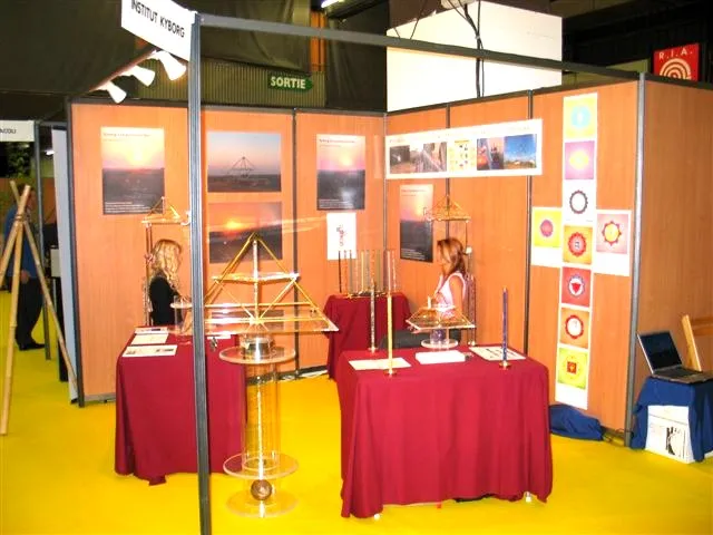 Messestand des Institut Kyborg auf dem Salon du Bien-Etre in Dijon: zwei Personen an mit bordeauxroten Tischdecken dekorierten Tischen, darauf mehrere Horus Energiepyramiden aus Metall; im Hintergrund Fotomontagen und Chakra-Grafiken an der Standwand.