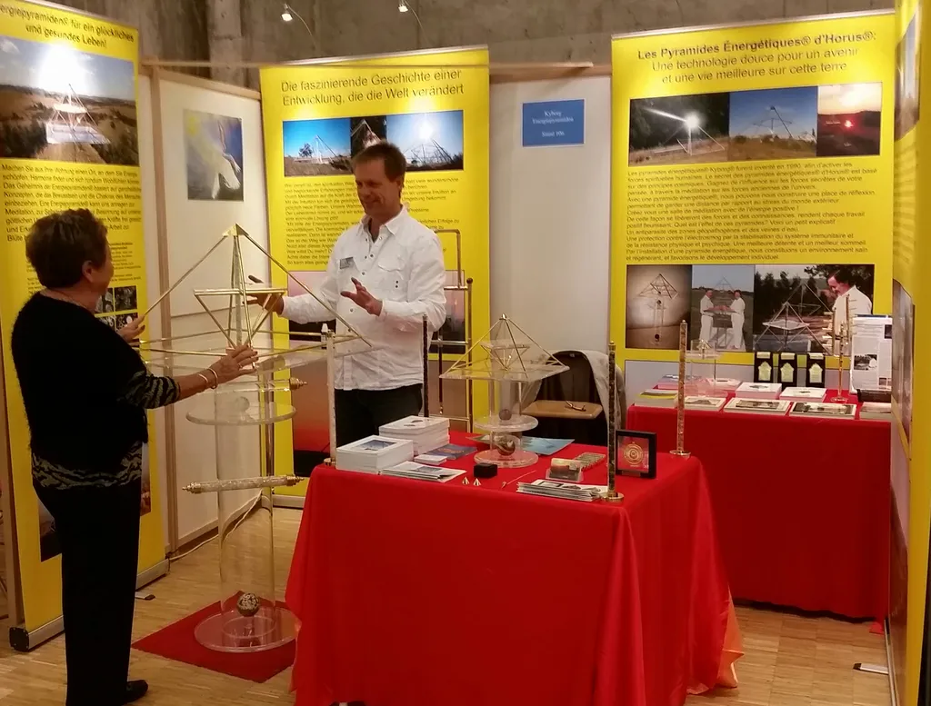 Mats Björkman im Gespräch mit einer Besucherin am Messestand des Kyborg Instituts mit Horus Energiepyramiden auf roten Tischen, umgeben von gelben Banner-Displays auf dem Salon de mieux vivre in Fribourg.