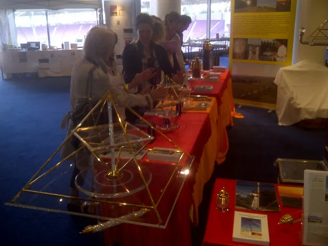 Messestand mit Horus Energiepyramiden aus Metall und Acrylglas auf einem rot gedeckten Tisch; zwei Besucherinnen betrachten die ausgestellten Objekte, im Hintergrund ein gelbes Informationsposter.