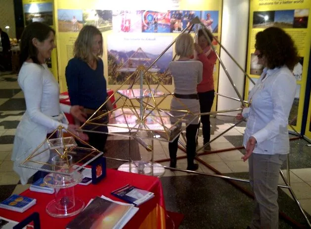 Messestand mit mehreren goldenen Horus Energiepyramiden unterschiedlicher Größe; vier Frauen besichtigen die Ausstellungsstücke vor einem gelben Standhintergrund mit Bildern und Texttafeln.