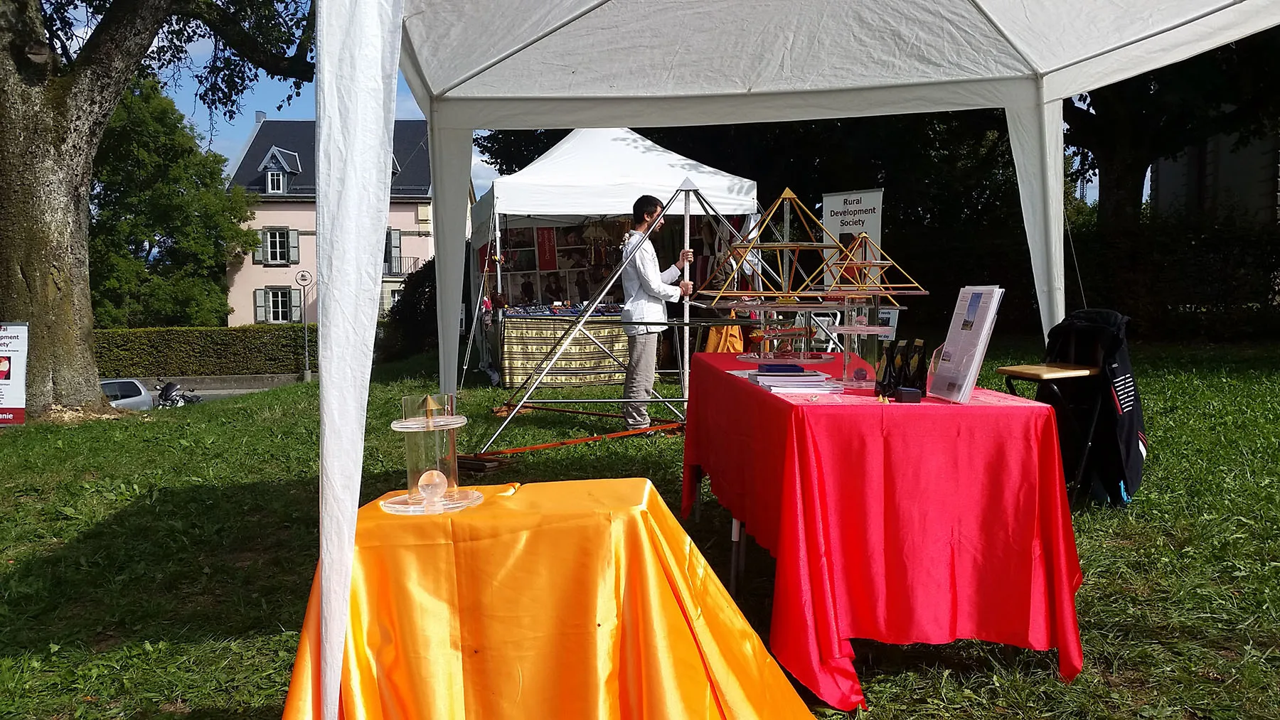 Messestand der Horus Energiepyramiden bei den Yogadays.ch in Assens: orangefarbener und roter Tisch mit Pyramiden-Modellen und Informationsmaterial unter einem weissen Pavillon im Freien, im Hintergrund eine weitere Pavillon-Einheit.