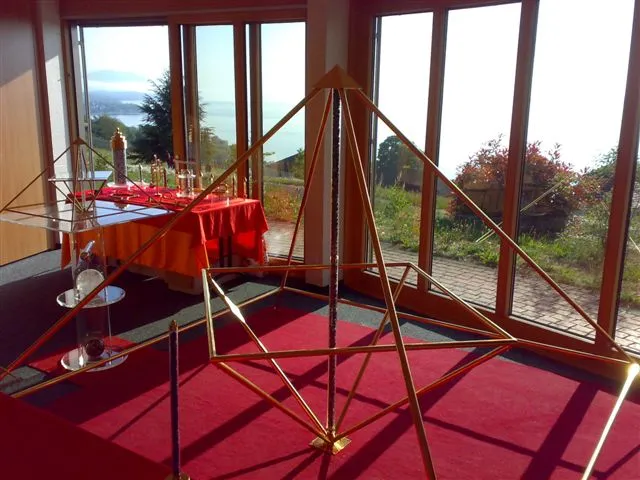Großes goldenes Horus-Energiepyramiden-Gestell aus Metallstäben in einem hellen Raum mit Panoramafenster und Seeblick; im Hintergrund ein rot gedeckter Tisch mit Ausstellungsobjekten.