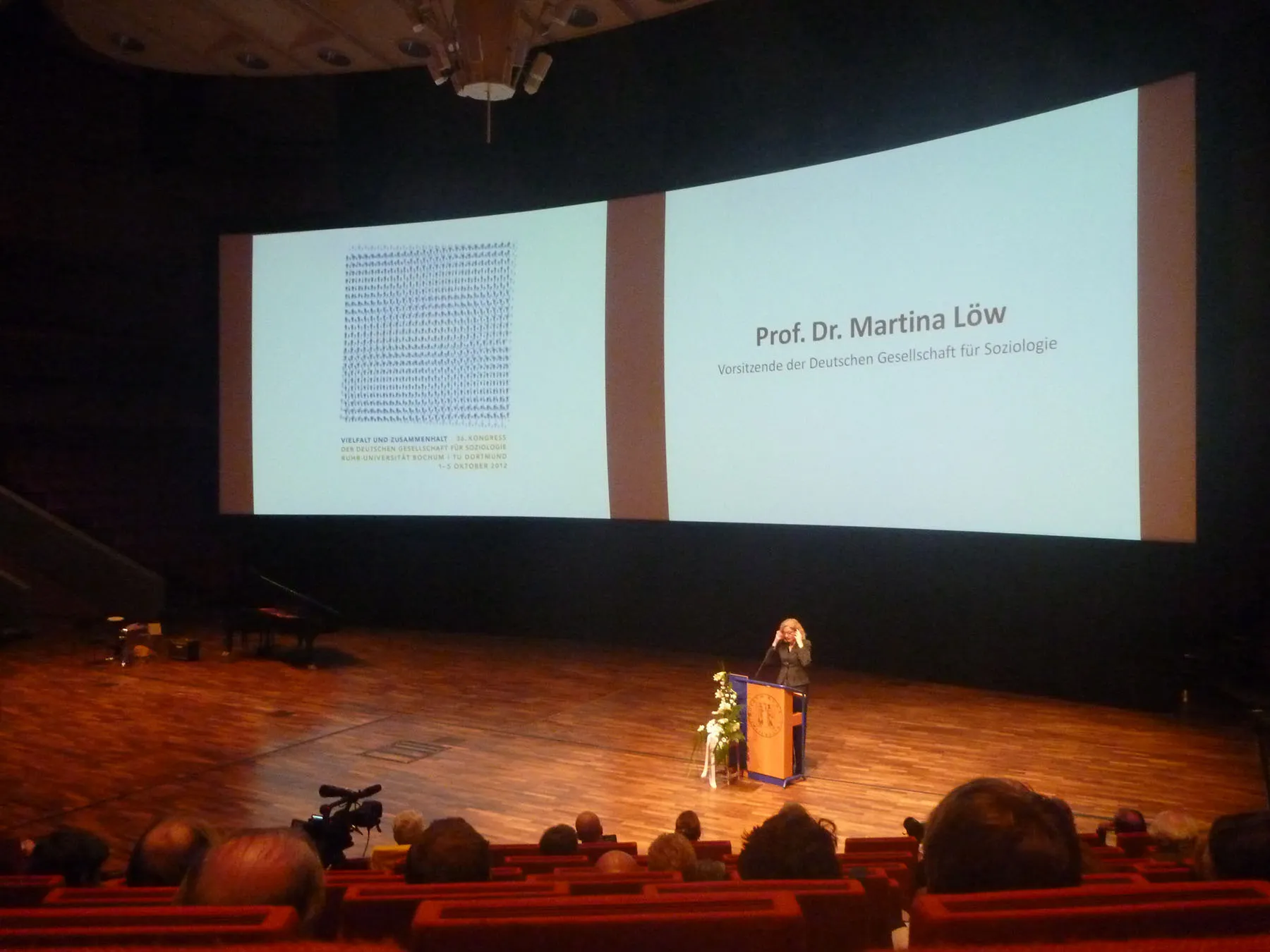 Eröffnungsveranstaltung des 36. DGS-Kongresses: Prof. Dr. Martina Löw spricht am Rednerpult in einem großen Auditorium, im Hintergrund die Leinwand mit Kongresstitel und Rednername.