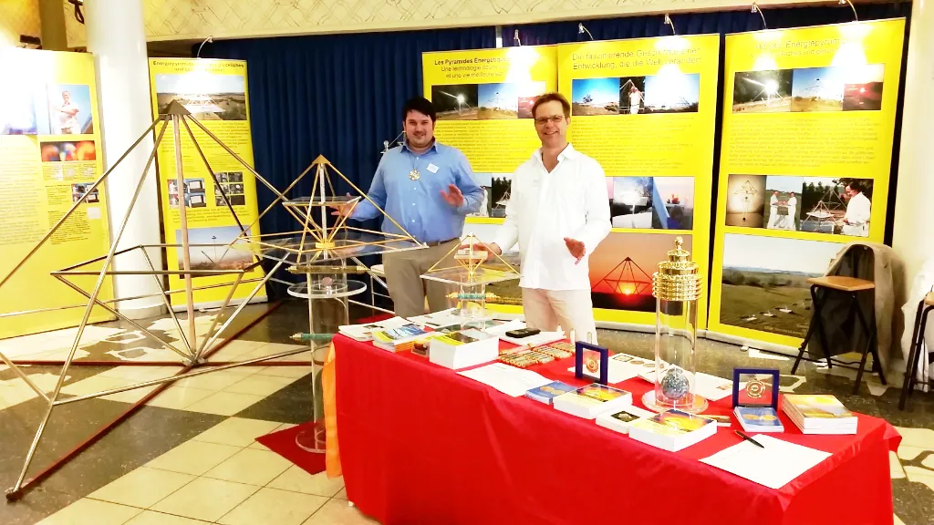 Zwei Männer stehen hinter einem rot gedeckten Messestand mit Horus Energiepyramiden und Büchern; im Hintergrund gelbe Aufsteller mit Produktfotos – Kyborg-Institut-Stand auf der Lebenskraft-Messe Zürich 2016.