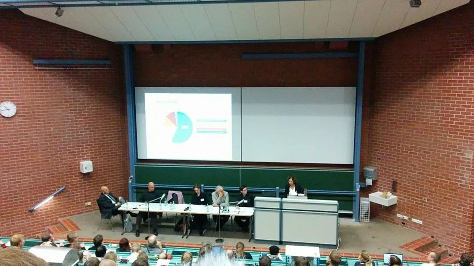 Blick von oben in einen Uni-Hörsaal mit Backsteinwänden: Sechs Personen auf dem Podium an einem langen Tisch, dahinter eine Leinwand mit einem Kuchendiagramm, davor ein gefülltes Auditorium.