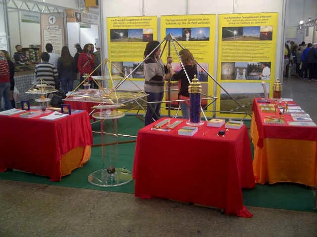 Messestand des Kyborg Instituts auf der Tisana in Lugano 2014: rot gedeckte Tische mit mehreren Horus Energiepyramiden aus Metall, dahinter dreisprachige Infotafeln (Deutsch, Französisch, Italienisch), zwei Besucherinnen im Gespräch am Stand.