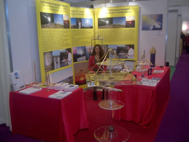 Messestand der Horus Energiepyramiden auf der Mednat 2014 in Lausanne: rote Tischdecken mit mehreren Pyramiden-Modellen aus Metall, gelbe Infotafeln im Hintergrund, eine Frau steht hinter dem Stand.