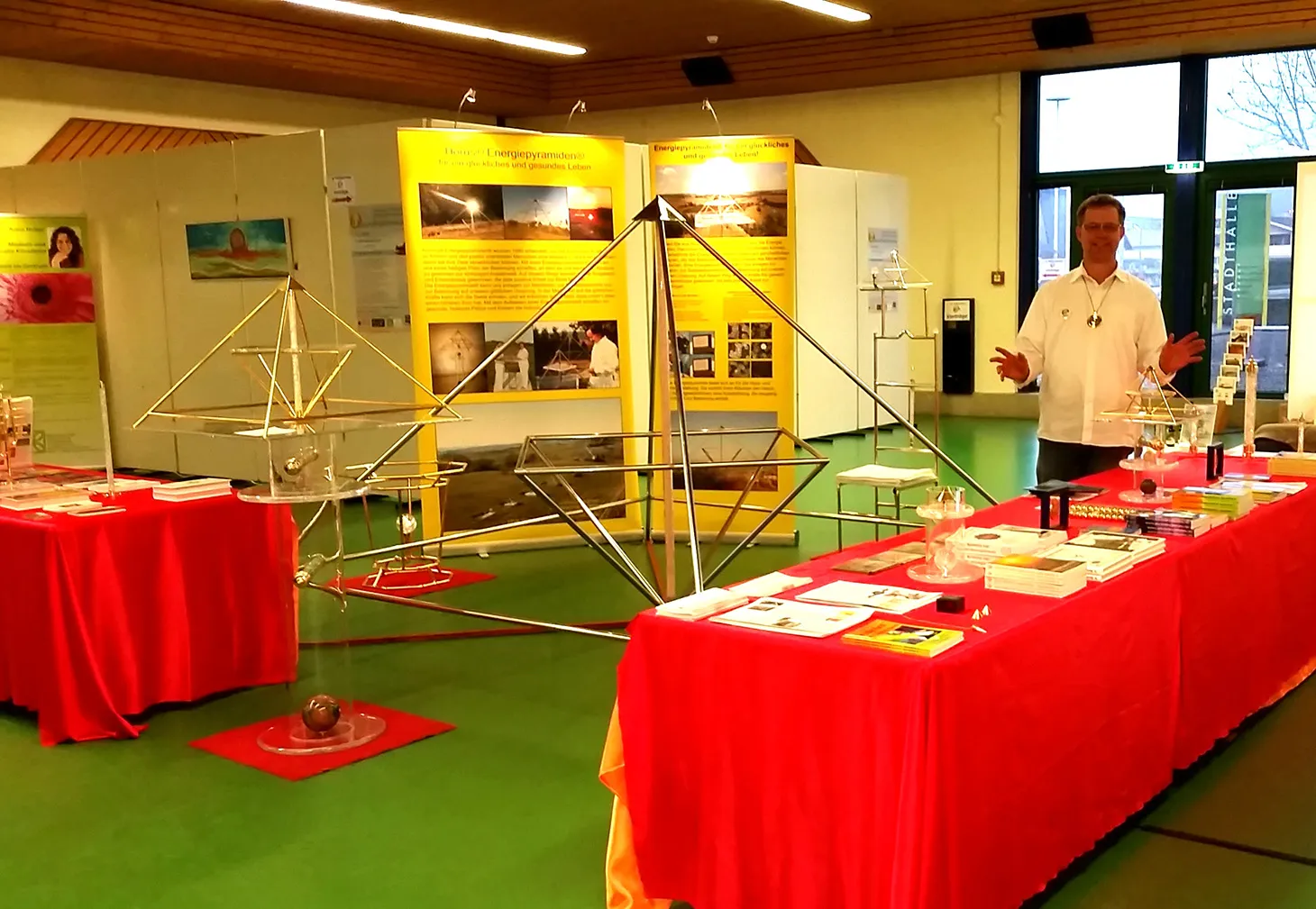 Mats Björkman steht an einem Messestand mit mehreren Horus Energiepyramiden aus Metall auf rot bezogenen Tischen sowie gelben Ausstellungsbannern des Kyborg Instituts im Hintergrund.