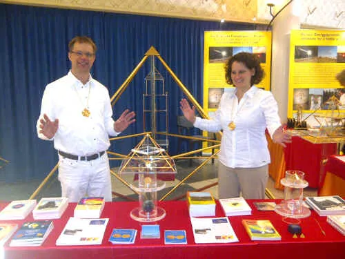 Mats und Ursula Björkman stehen lächelnd hinter einem rot gedeckten Messestand mit einer Horus Energiepyramide aus Messing und ausgelegten Informationsbroschüren; im Hintergrund gelbe Ausstellungstafeln.
