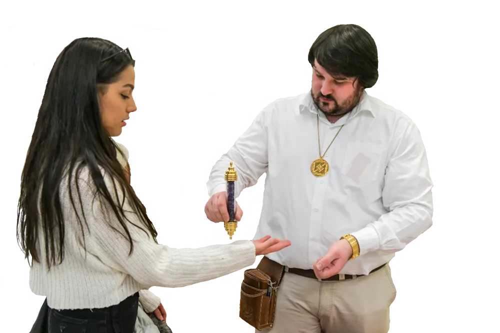 Tobias O. R. Alke im weißen Hemd mit Amulett hält den goldverzierten Powerstab B über die ausgestreckte Hand einer jungen Frau mit langen dunklen Haaren; dunkler Hintergrund (Energieübung bei einem Pressetermin).