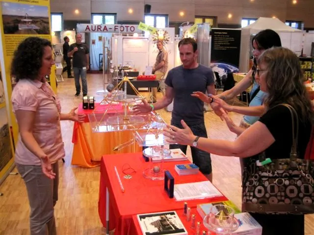 Ursula Björkman in rosa Bluse im Gespräch mit Besucherinnen und Besuchern an einem Messestand mit goldenen Energiepyramiden und Kristallständern auf rot-orange gedeckten Tischen.