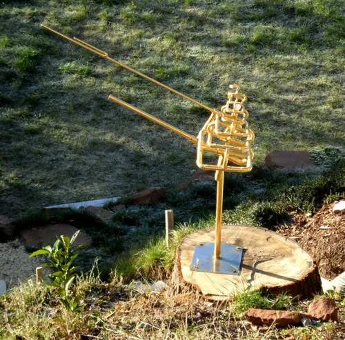 Vergoldete Skulptur Vidaya aus gebogenen Rohren: ein turmartig verschlungener Kopfteil mit zwei langen, schräg nach oben ragenden Rohren auf einem schlanken Stab, verschraubt auf einer Metallplatte auf einem Baumstumpf; dahinter reifbedeckter Rasen und Sandsteinbrocken.