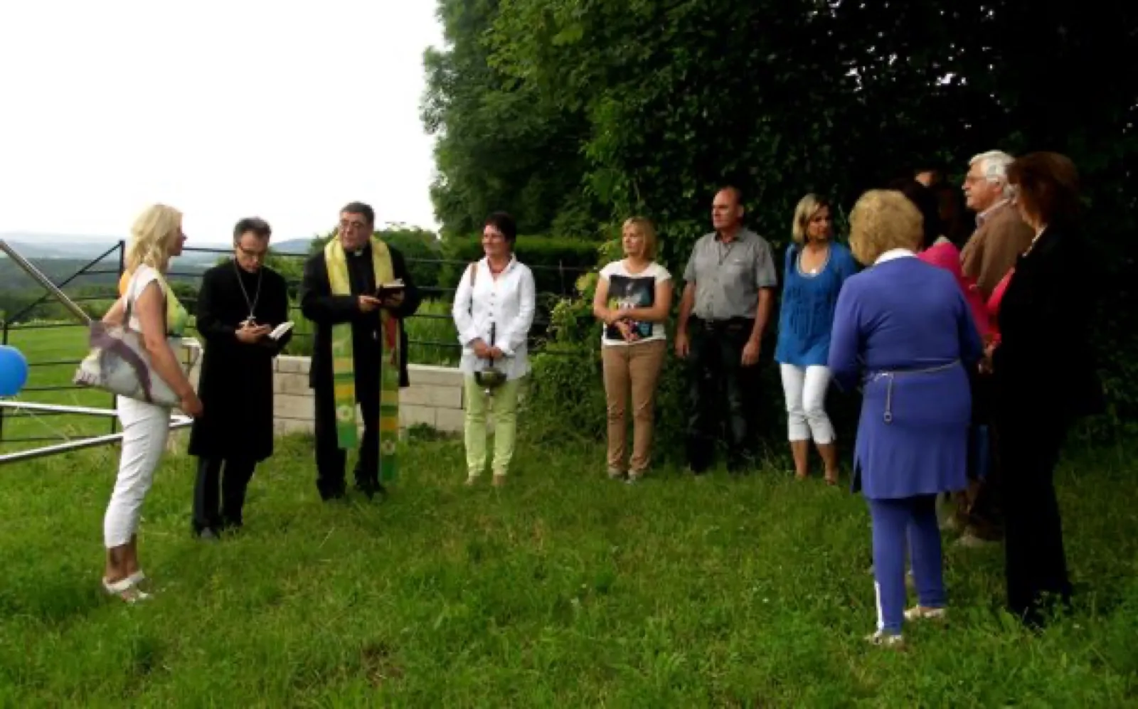Einweihung im Freien: Zwei Geistliche in schwarzer Kleidung, einer mit grün-gelber Stola und aufgeschlagenem Buch, stehen vor einer Gruppe von etwa zehn Menschen im Halbkreis auf einer Wiese, dahinter Bäume und ein Geländer.