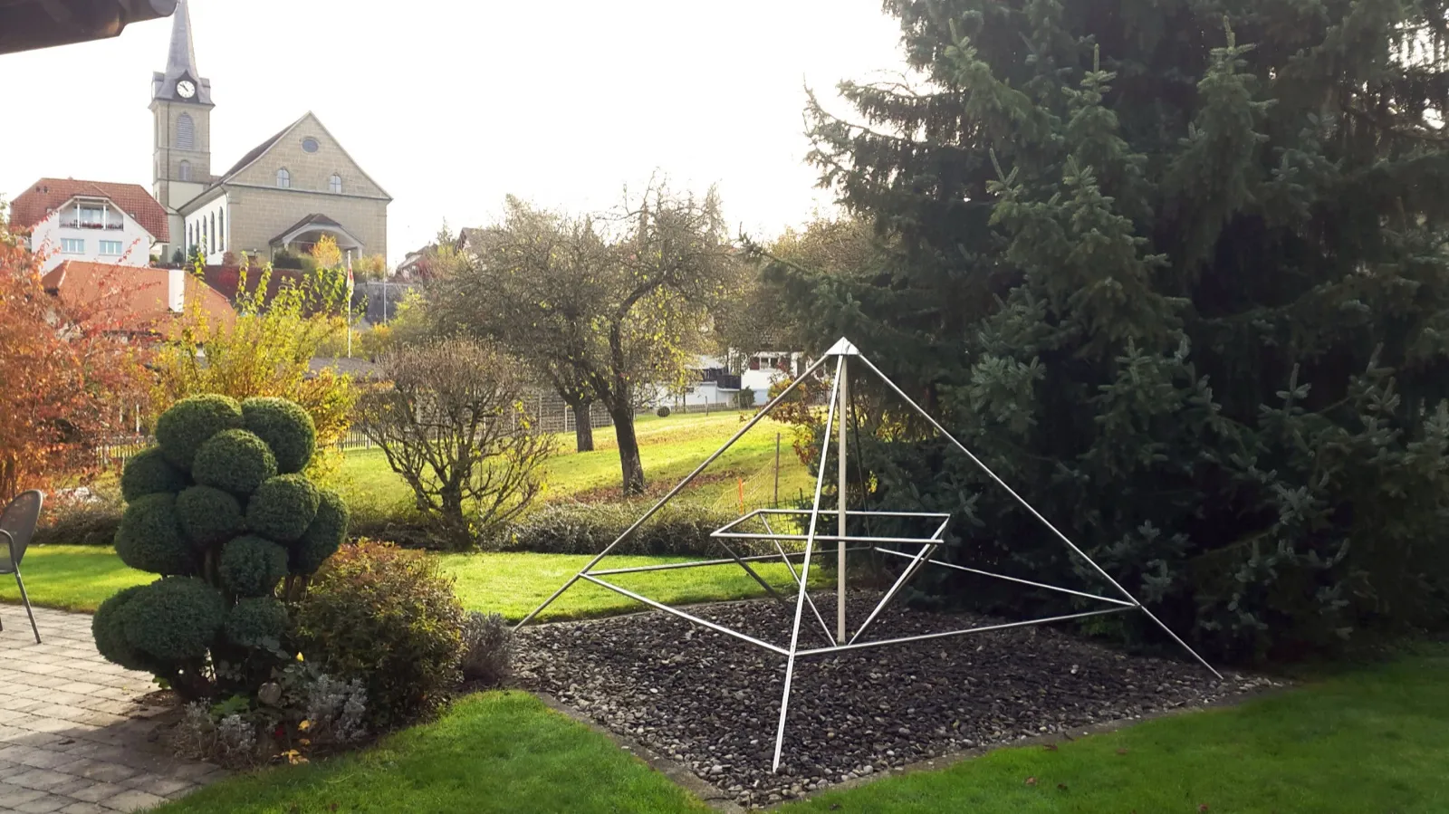 Offene Energiepyramide aus Edelstahlrohren auf einem Kiesbett in einem herbstlichen Garten, links ein kugelig geschnittener Buchsbaum und eine Terrasse, rechts eine große Fichte; im Hintergrund eine Kirche mit Uhrturm und Wohnhäuser.