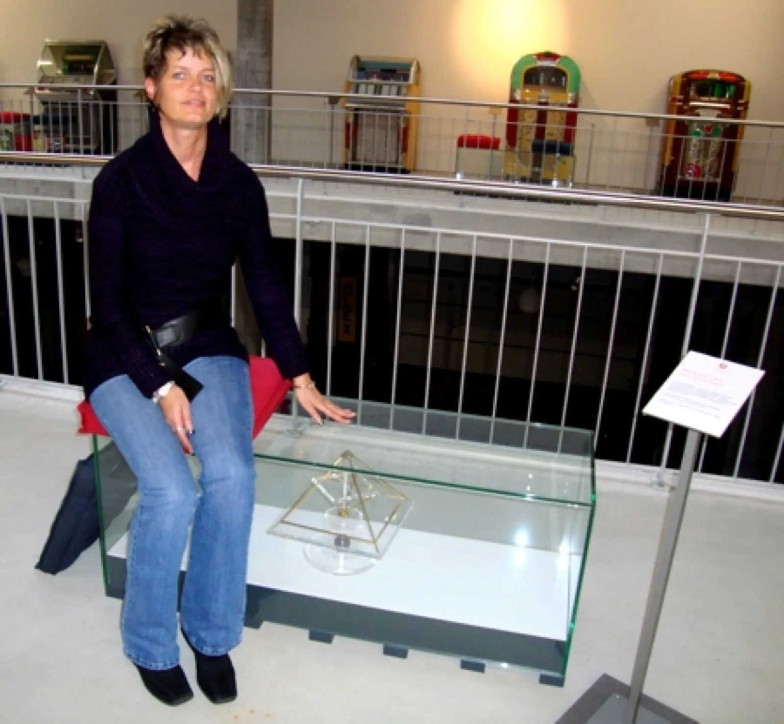 Eine Frau mit kurzem blondem Haar sitzt in einem Museum neben einer Glasvitrine, in der eine kleine Energiepyramide mit Acrylglasplatte steht; daneben ein Hinweisschild, im Hintergrund auf der Galerie mehrere historische Jukeboxen.