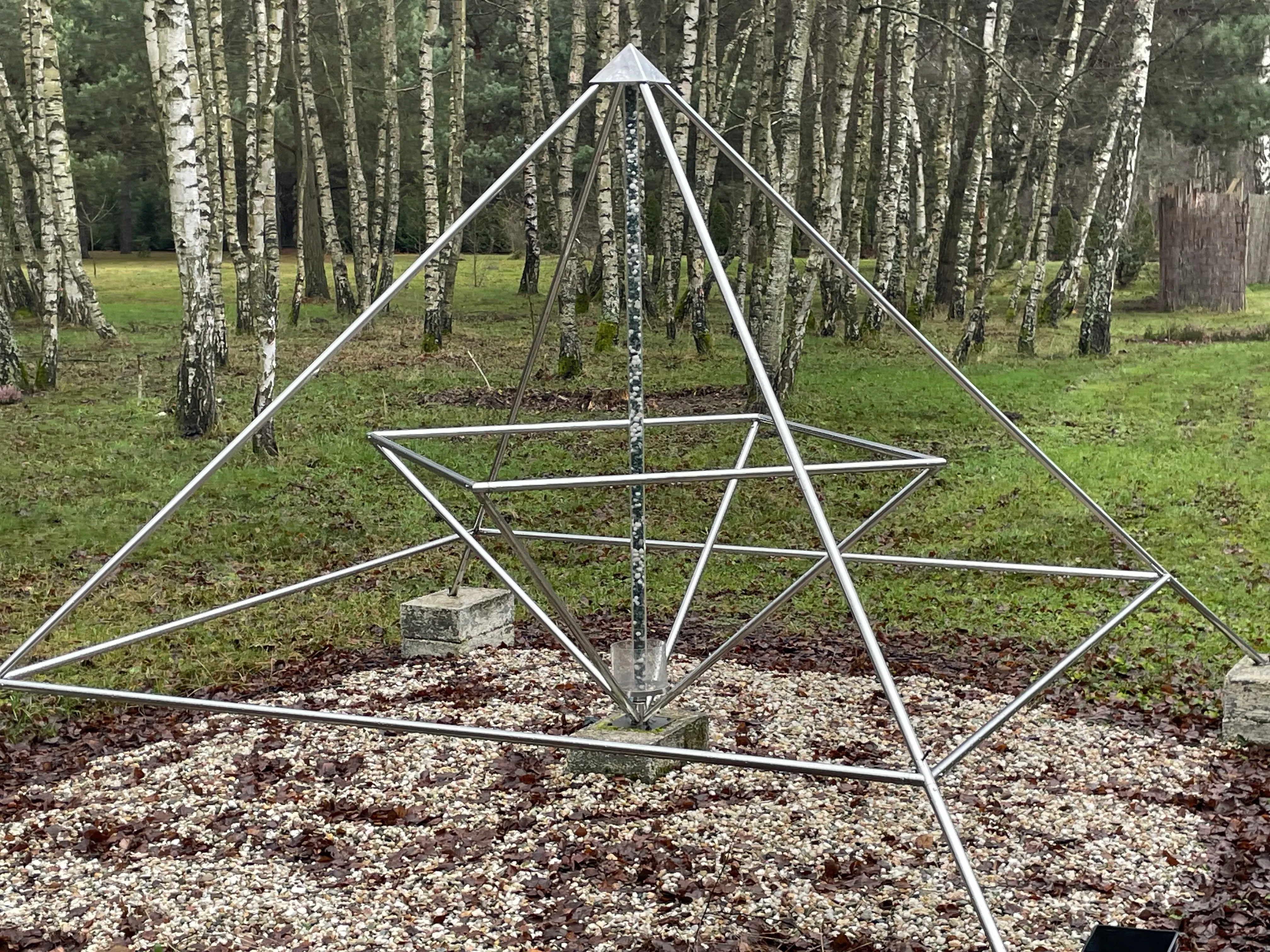 Energiepyramide Modell C als Edelstahl-Doppelpyramide zwischen Birken in einem naturnahen Garten; gut sichtbar der innere 45°-Stern, die zentrale Achse und die vier Füße auf Steinsockeln.