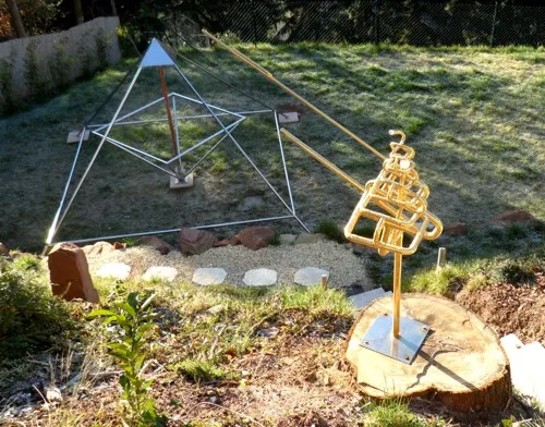 Übersicht über den Garten in Wintersonne: links eine offene Energiepyramide mit kupferfarbener Mittelachse, rechts hinten eine zweite große Pyramide, in der eine Person in blauer Jacke steht, in der Mitte die vergoldete Skulptur Vidaya auf einem Baumstumpf. Dahinter ein Maschendrahtzaun und dunkler Nadelwald am Hang.