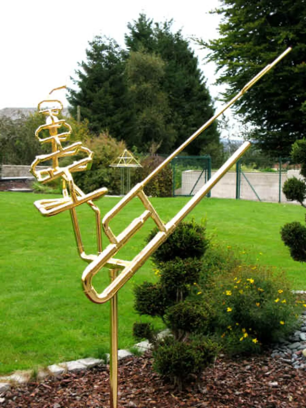 Eine hohe goldglänzende Skulptur aus Rohren in Form einer stilisierten Figur mit langem Stab steht in einem Gartenbeet mit Formgehölzen; im Hintergrund auf dem Rasen eine kleine goldfarbene Pyramide vor Nadelbäumen und Zaun.