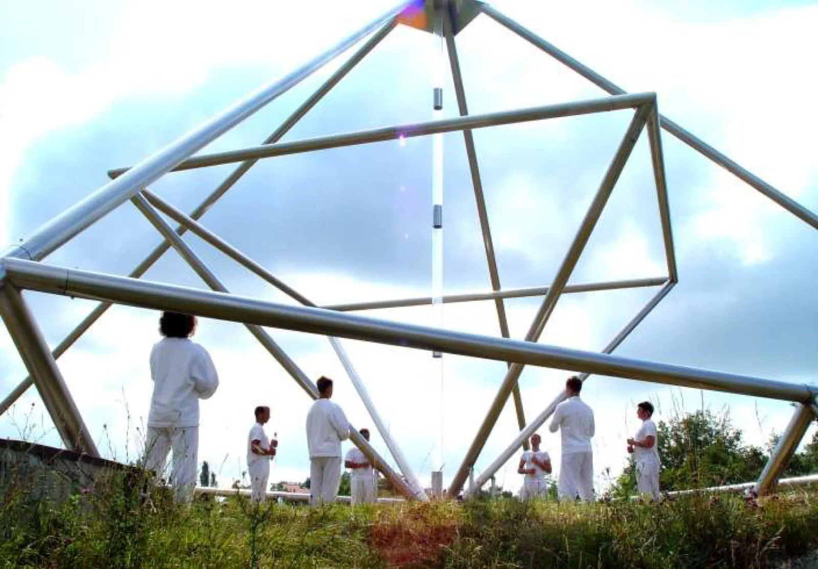 Blick von unten in eine große offene Pyramide aus silbernen Metallrohren mit mittiger durchsichtiger Achse; darunter stehen sieben Menschen in weißer Kleidung verteilt im hohen Gras vor bewölktem Himmel.