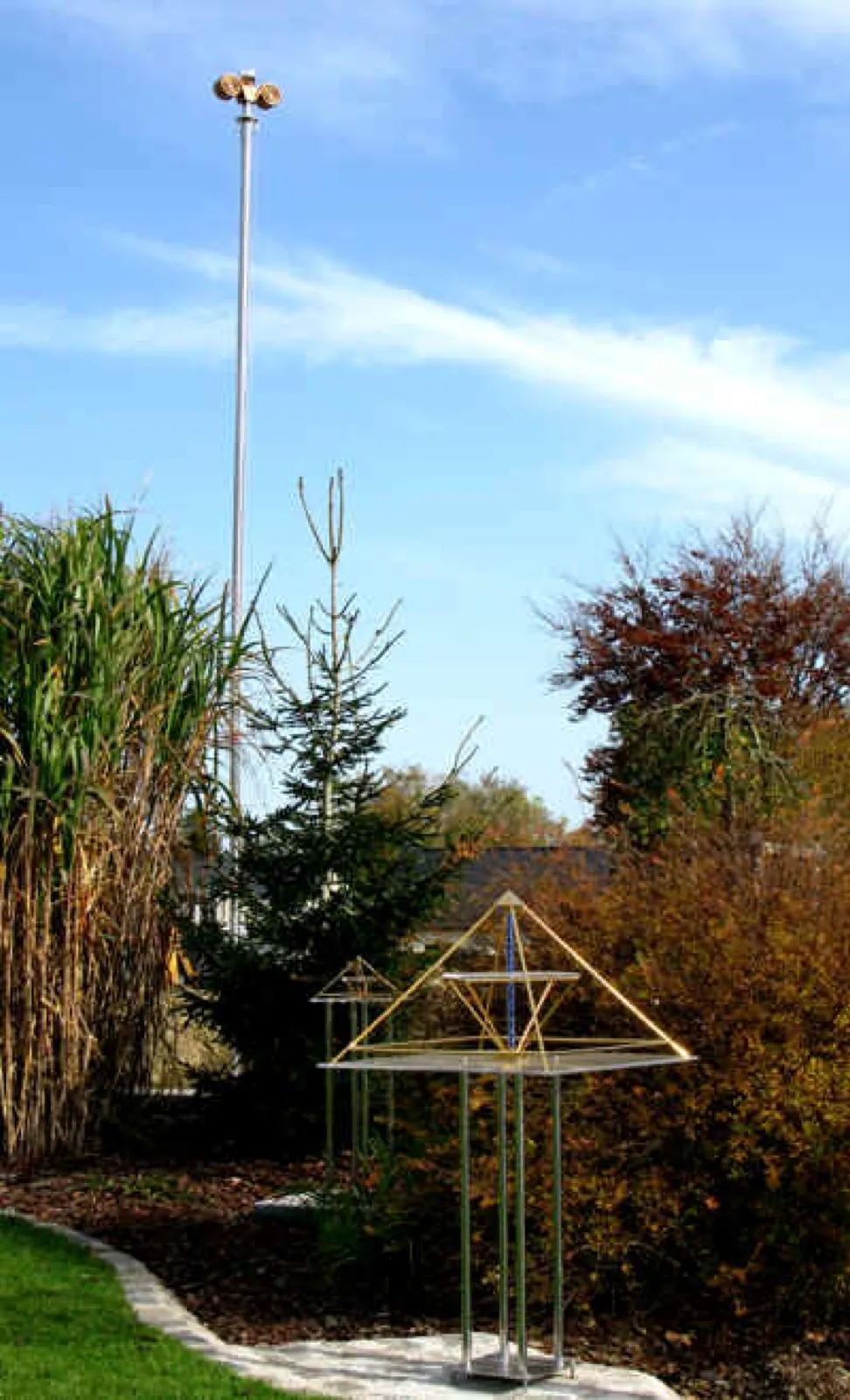 Ein hoher schlanker Metallmast mit goldfarbenem, spiralförmigem Aufsatz ragt in den blauen Himmel; davor stehen zwei goldfarbene Energiepyramiden auf Edelstahlgestellen zwischen herbstlich gefärbten Sträuchern, Chinaschilf und einer kleinen Fichte.