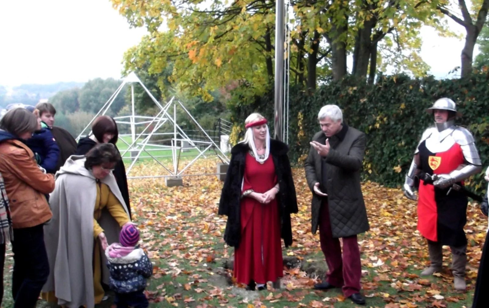 Kleine Zeremonie auf herbstlichem Laub: Eine Frau in rotem mittelalterlichem Kleid mit schwarzem Umhang und ein grauhaariger Mann im dunklen Mantel stehen an einem Mast, rechts ein Mann in Ritterrüstung mit Helm; links Zuschauer, dahinter eine offene Metallpyramide.