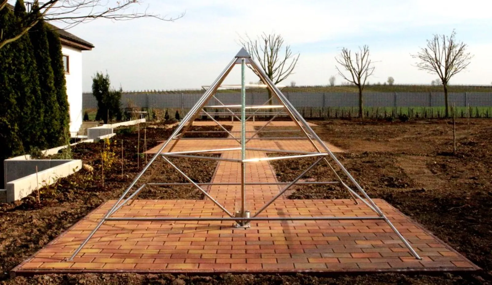 Offene Energiepyramide aus Edelstahlrohren mit Mittelachse auf einer Fläche aus rötlichem Klinkerpflaster, dahinter ein gepflasterter Weg zu weiteren Pyramidengestellen; ringsum frisch angelegte Beete, junge kahle Bäume, ein weißes Haus und ein Zaun vor Weinbergen.