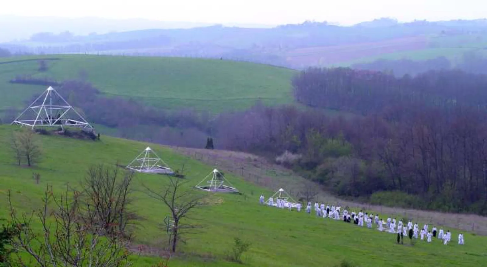 Hügelige, dunstige Frühlingslandschaft von Le Braquet: Auf einem grünen Hang stehen vier offene Pyramidengestelle unterschiedlicher Größe, neben dem kleinsten übt eine Gruppe in weißer Kleidung in Reihen.