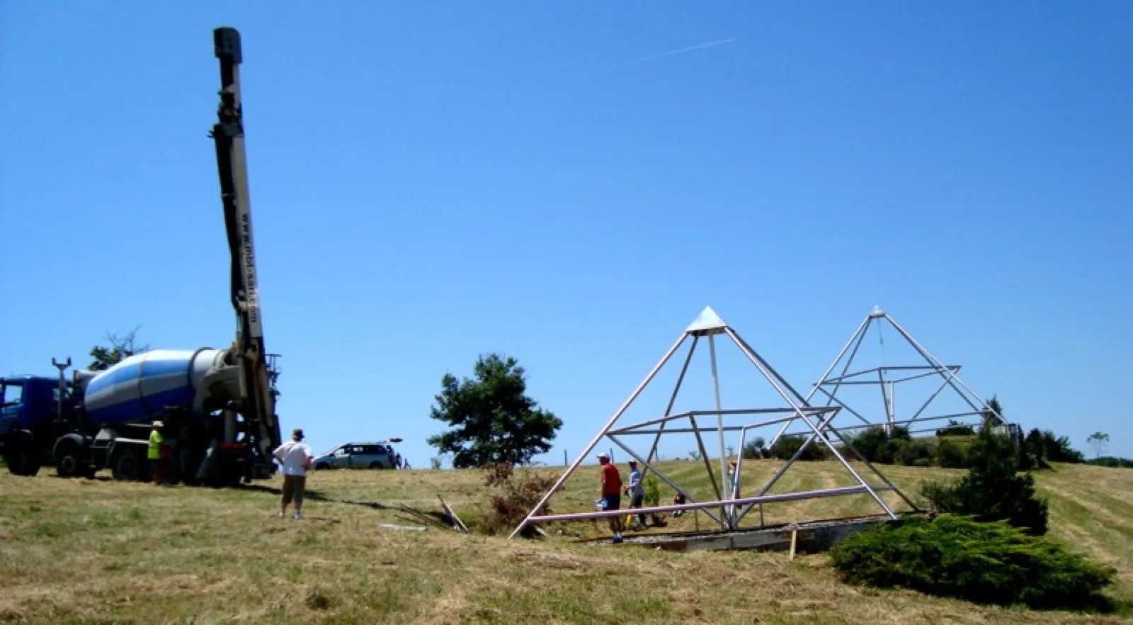 Auf einer sonnigen Hügelwiese steht links ein Betonmischer-Lkw mit ausgefahrenem Pumpenmast, rechts zwei große offene Pyramidengestelle aus Metall, an denen mehrere Menschen arbeiten. Blauer Himmel, ein Baum und ein geparktes Auto im Hintergrund.
