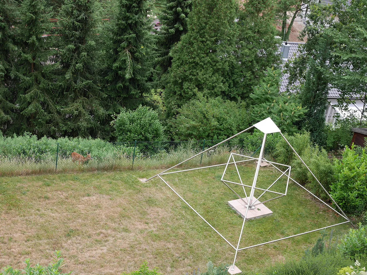 Derselbe Garten aus gleicher Perspektive: Hinter dem Maschendrahtzaun am Wiesenrand steht ein einzelnes Reh im hohen Gras; rechts die offene Edelstahl-Energiepyramide mit innenliegendem Oktaeder, im Hintergrund Nadelbäume und ein graues Hausdach.
