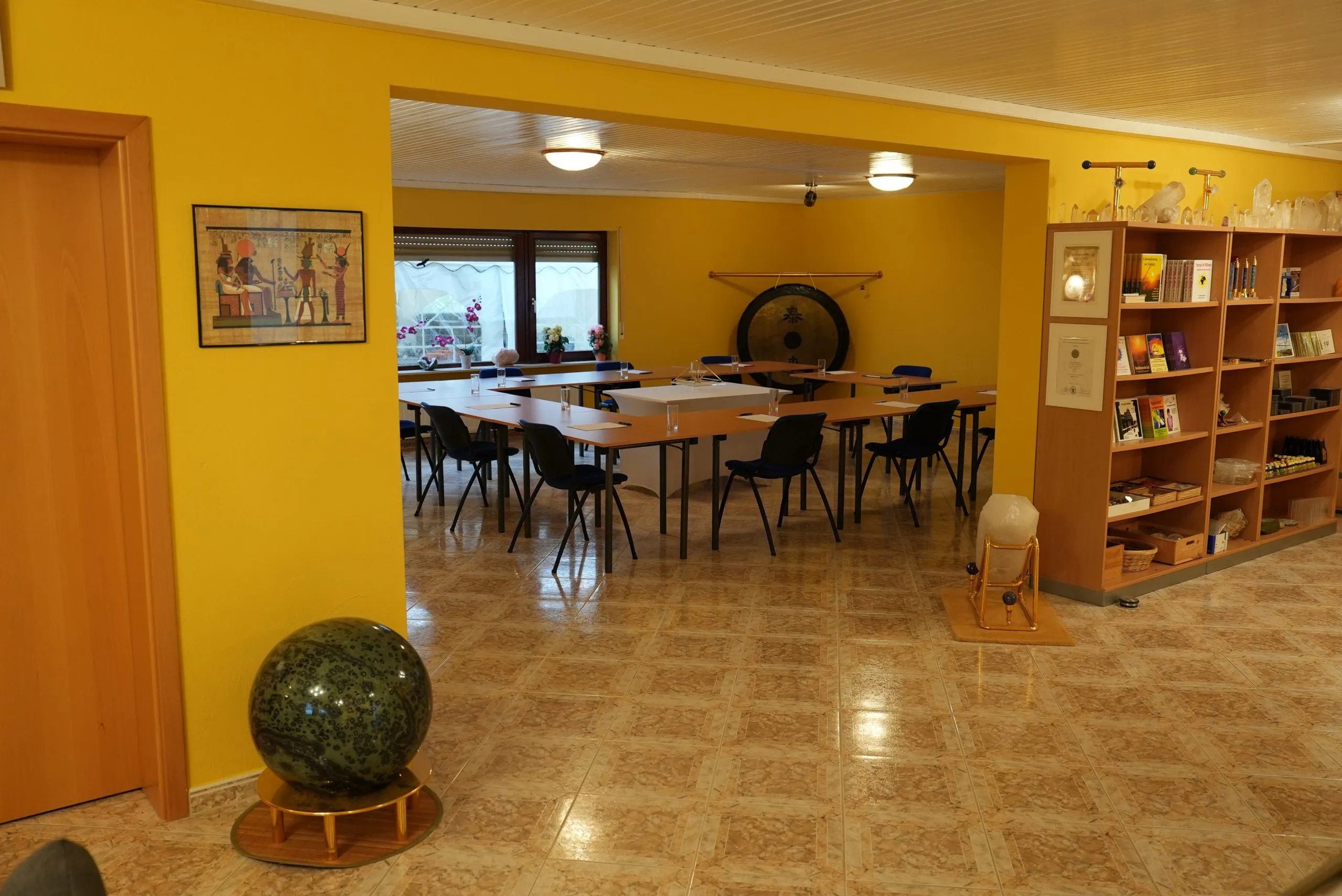 Heller Seminarraum mit langer Tischreihe und Stühlen, einem großen Gong, einem ägyptischen Papyrus-Bild an der Wand und einer Bücherwand mit Kristallkugeln.