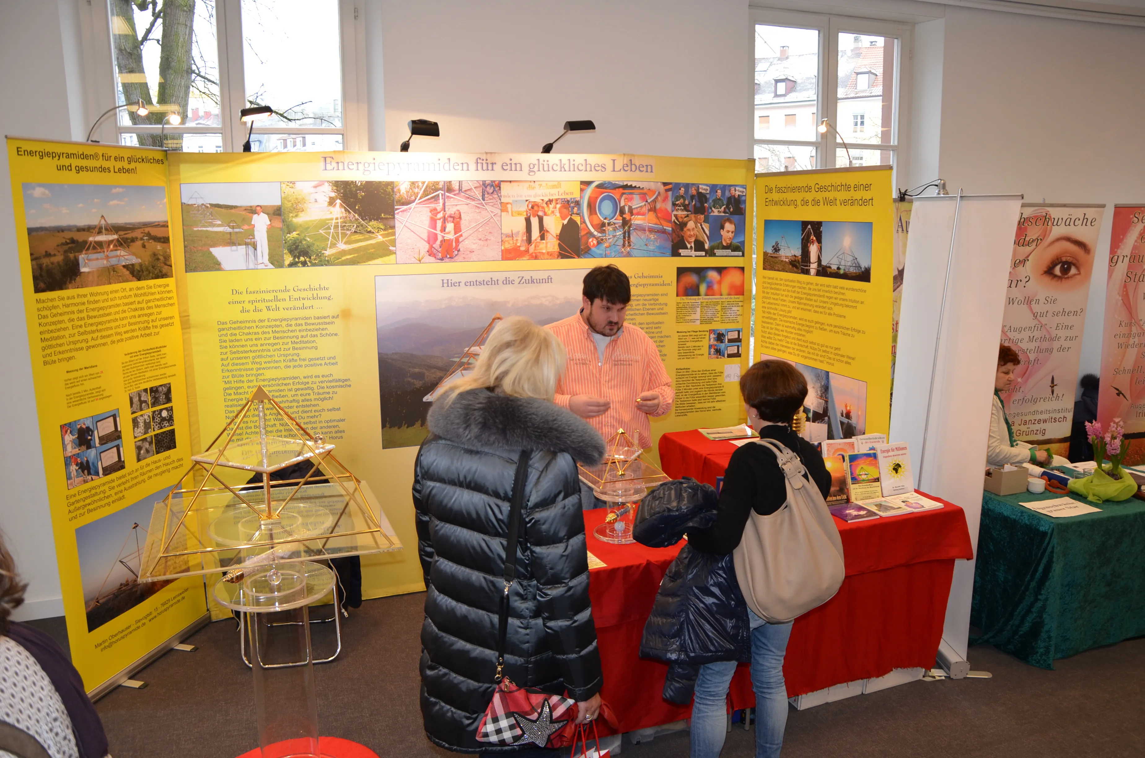 Tobias Alke hinter dem Messestand im Gespräch mit zwei Besucherinnen; gelbe Infowand „Energiepyramiden für ein glückliches Leben“, ein goldenes Pyramidenmodell auf einer Acrylsäule und ein rot gedeckter Tisch mit Büchern und Broschüren.