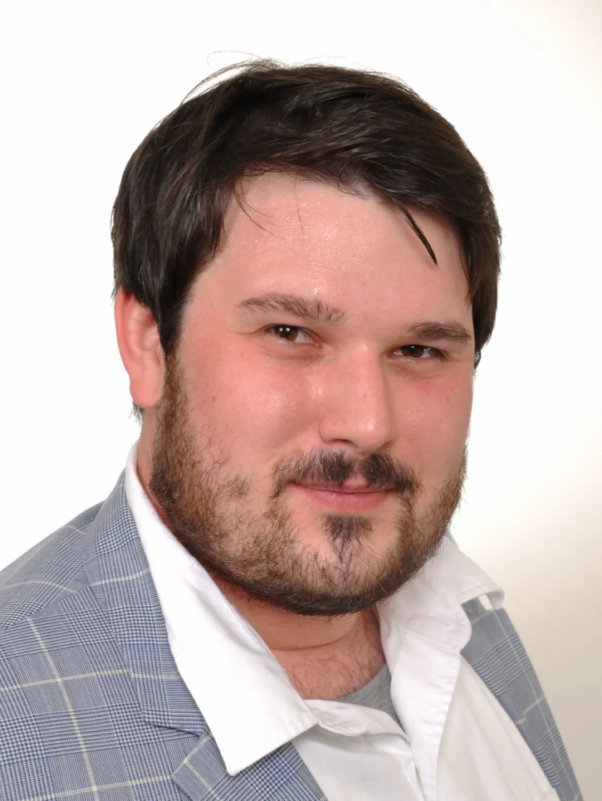 Porträt von Tobias O. R. Alke mit dunklem Haar und Vollbart, in weißem Hemd und hellblau kariertem Sakko vor hellem Hintergrund.