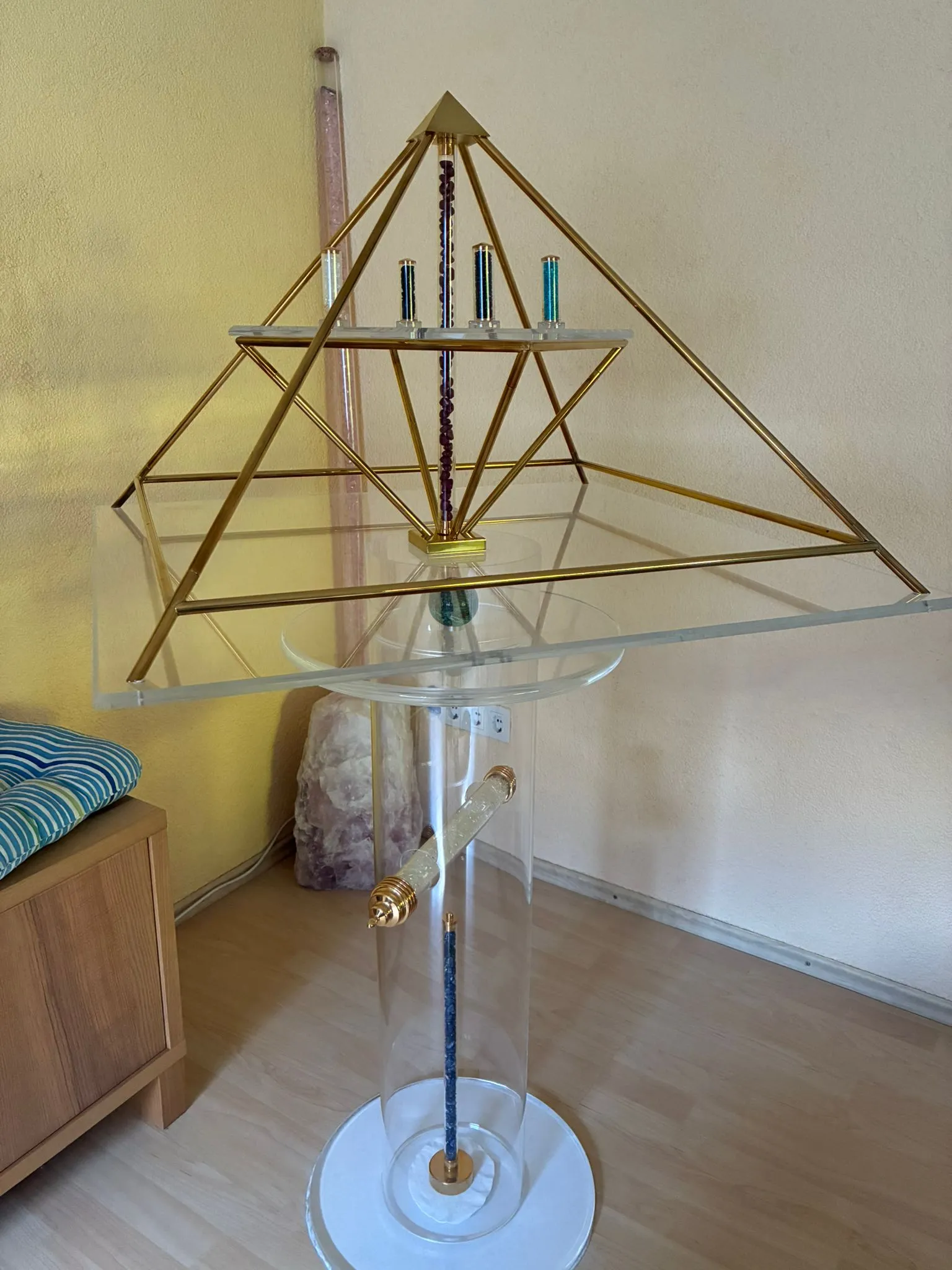 Vergoldete Energiepyramide auf einer durchsichtigen Acrylsäule, im Inneren Edelstein-Stäbe (violetter Amethyst, blaue und grüne Röhren); am Sockel eine Amethyst-Druse, daneben eine Sitzbank.