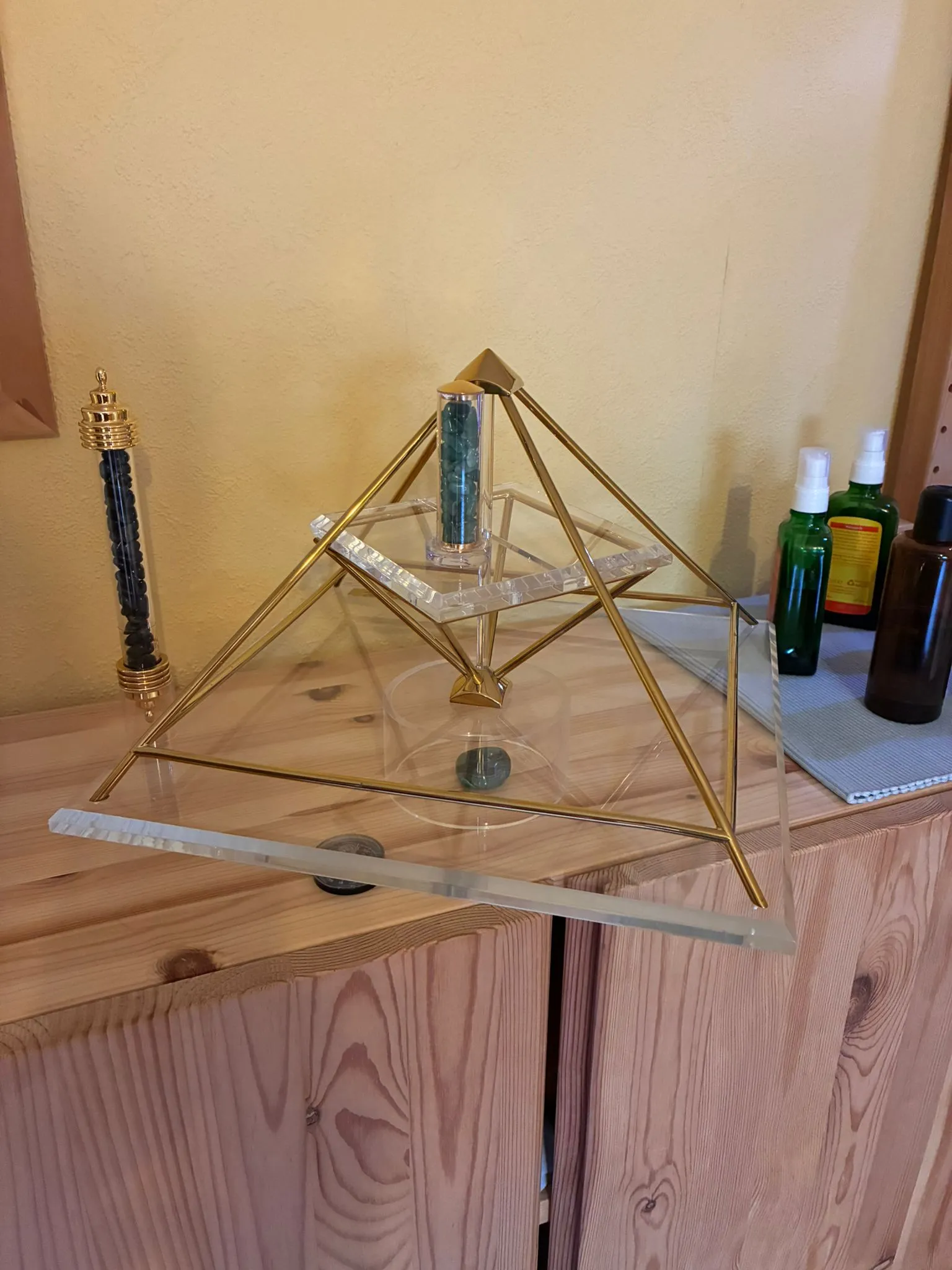 Vergoldete Energiepyramide Modell A auf einem Holzschrank mit grünem Edelstein-Stab; links ein Stab mit schwarzen Steinen, rechts kleine Aromaöl-Fläschchen.