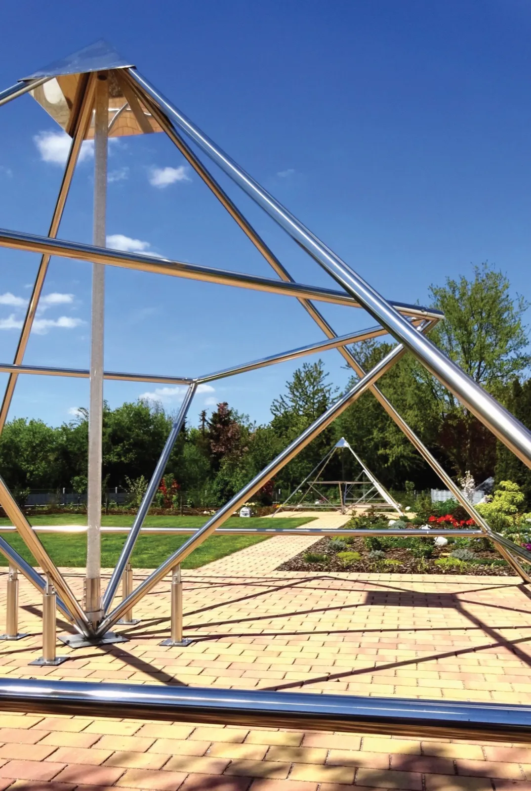 Nahansicht einer großen, spiegelnd polierten Edelstahlpyramide mit Spitzenblech und heller Mittelachse auf gepflasterter Fläche unter tiefblauem Himmel. Durch das Gestell blickt man auf einen Gartenweg, Blumenbeete mit roten Blüten und eine weitere Pyramide im Hintergrund.