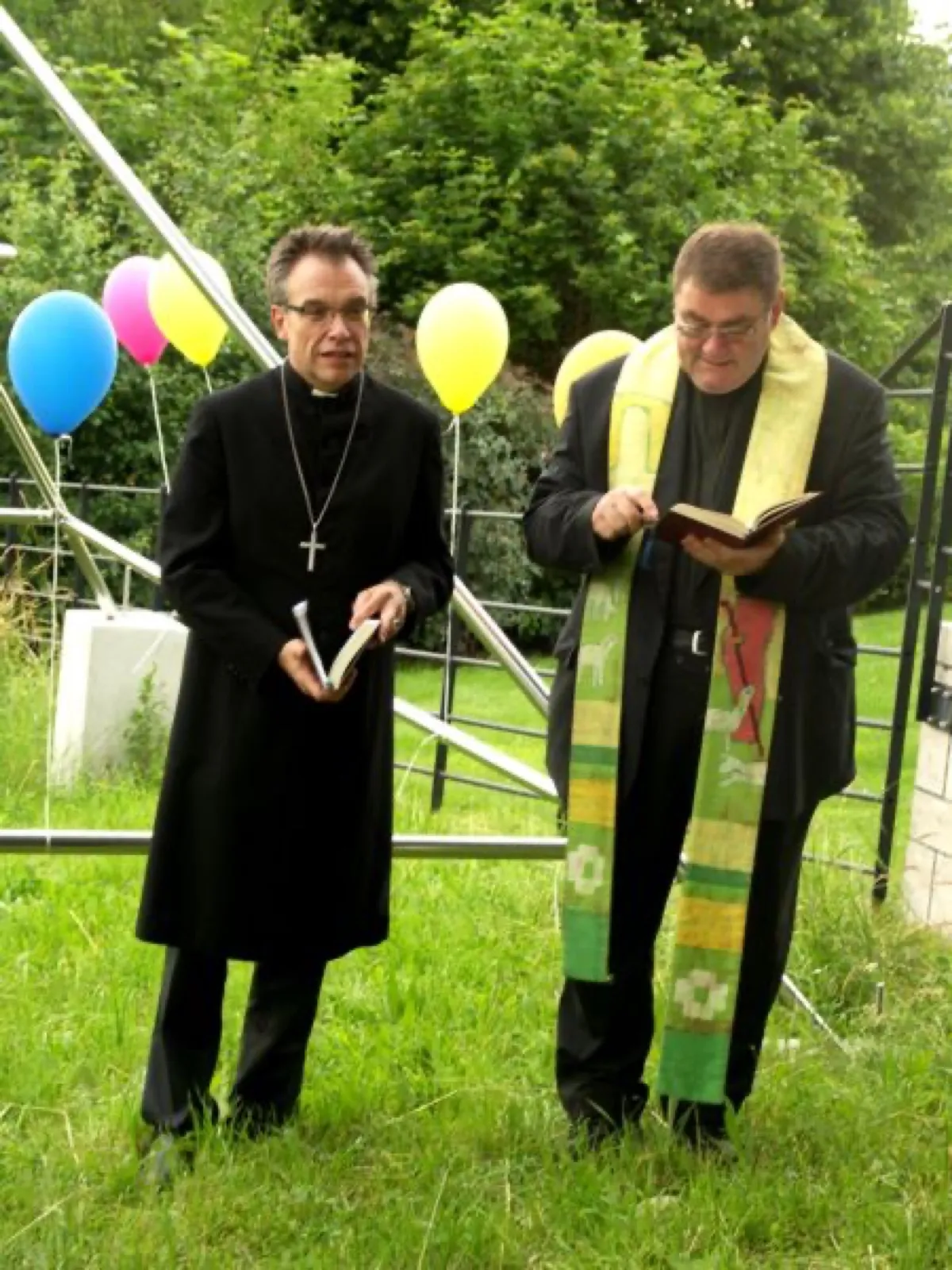 Zwei Geistliche stehen auf einer Wiese vor einem Pyramidengestell aus Metallrohren: links ein Mann in schwarzem Talar mit Brustkreuz und Buch, rechts ein Mann mit grün-gelber Stola, der aus einem Buch liest; dahinter bunte Luftballons, ein Zaun und Büsche.